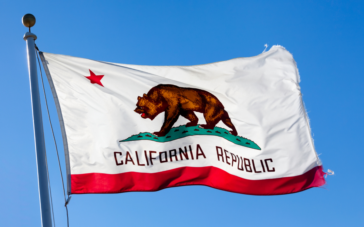 The California Republic flag flies against a clear blue sky. The flag features a brown bear walking on a green patch of grass, a red star in the upper left corner, and the words "CALIFORNIA REPUBLIC" written below the bear. A red stripe runs along the bottom edge of the flag.