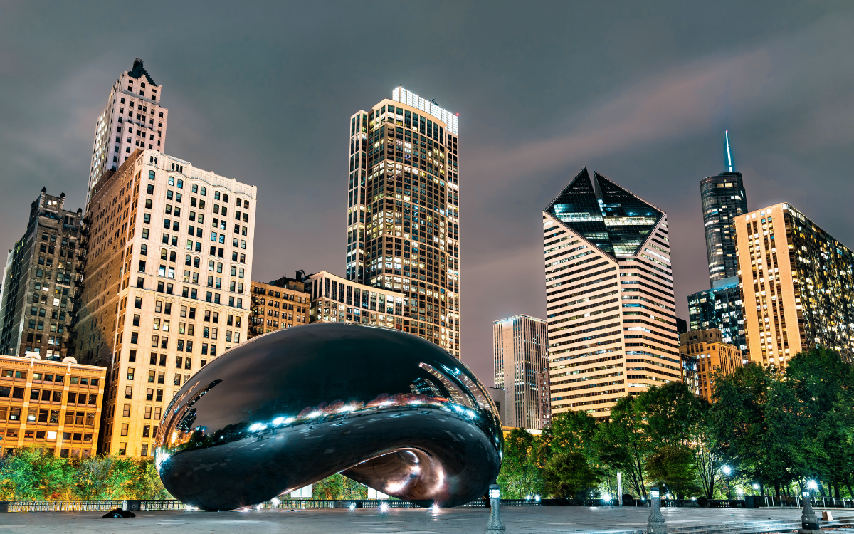 The Cloud Gate sculpture reflects the Chicago skyline at night under a cloudy sky. Tall skyscrapers with illuminated windows tower behind the large, reflective bean-shaped art installation.