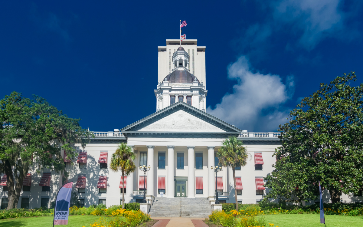 The Florida State Capitol building in Tallahassee, with a bright blue sky and white clouds overhead. The building is white with columns and a large dome, surrounded by green lawns and trees.