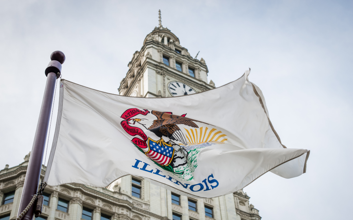 The Illinois state flag, featuring the state seal with an eagle and the word "ILLINOIS", waves in front of a building with a clock tower.