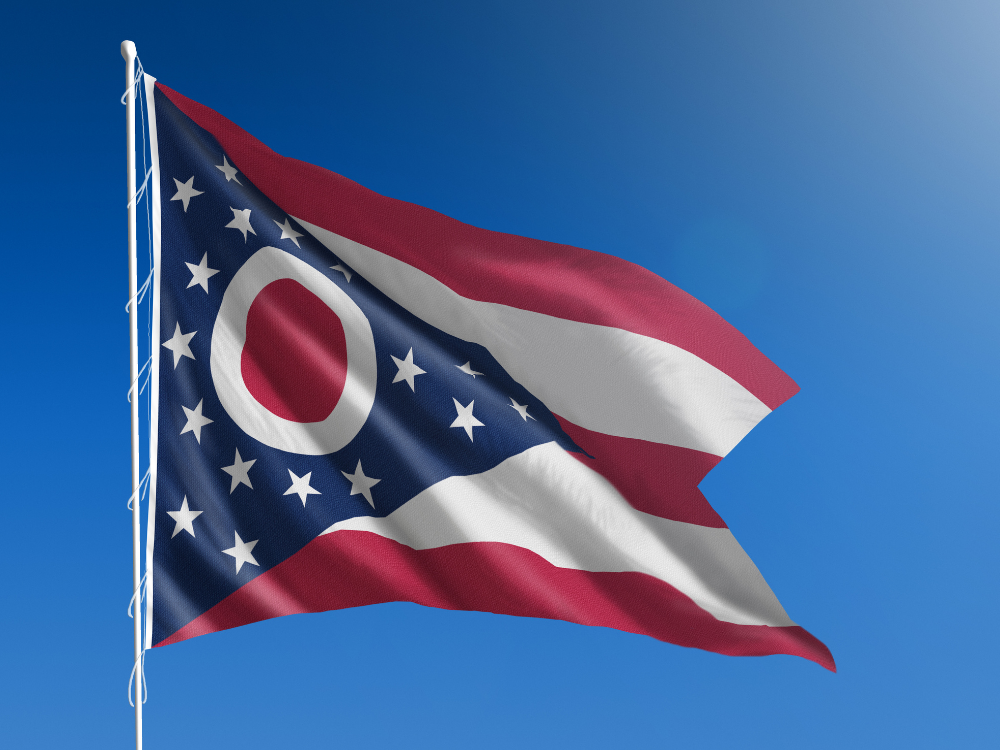 The Ohio state flag, a swallowtail pennant with red and white stripes, a blue triangle, and a white circle with a red center surrounded by 13 white stars, waves against a clear blue sky.