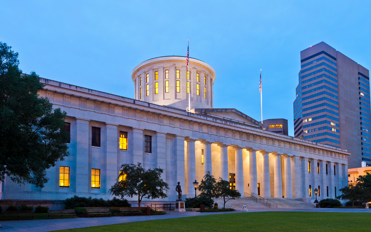 The Ohio Statehouse with its dome illuminated against a twilight sky, flanked by trees and a modern skyscraper.