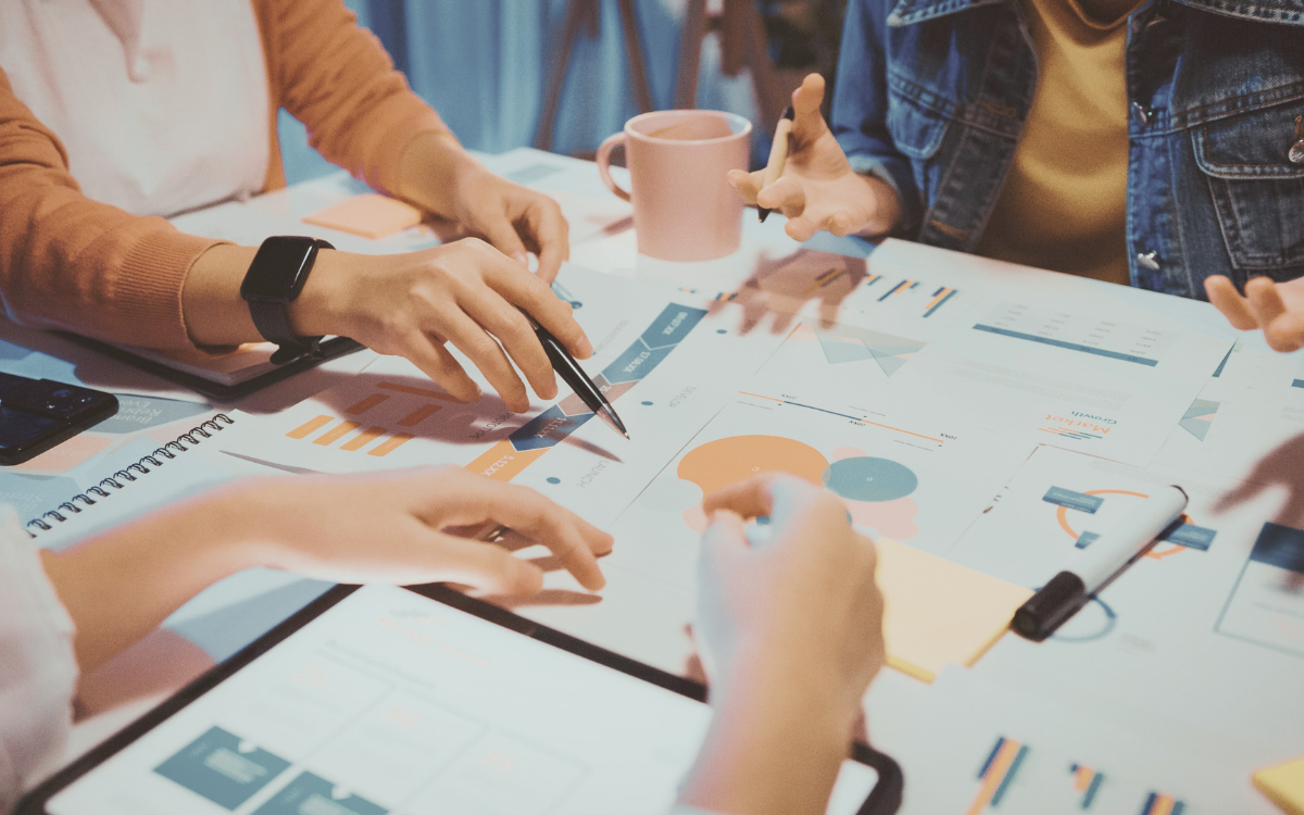 A group of people gathered around a table, pointing at charts and graphs on papers, discussing business strategies.