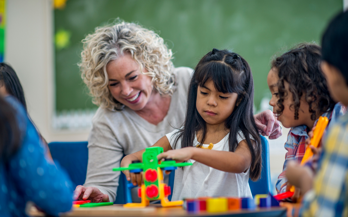 A teacher helps a child build a toy structure at a table in a classroom.