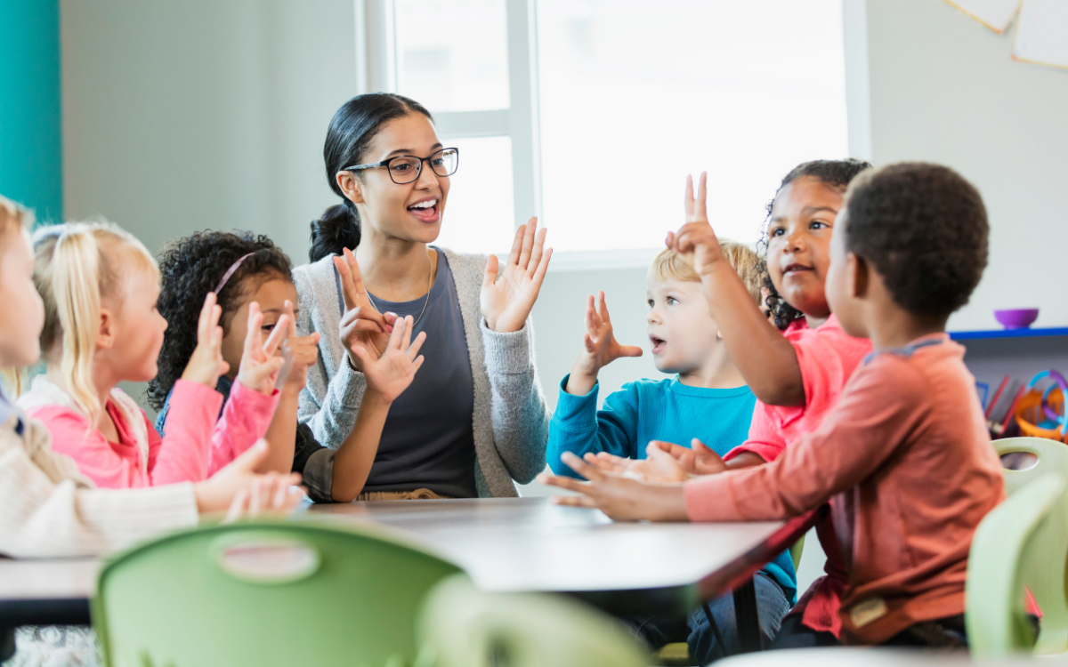A teacher with glasses leads a group of young children in a classroom activity, with everyone raising their hands.