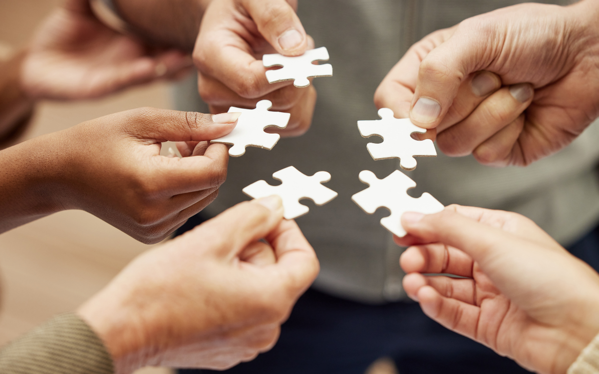 A group of hands holding several white puzzle pieces, attempting to connect them.